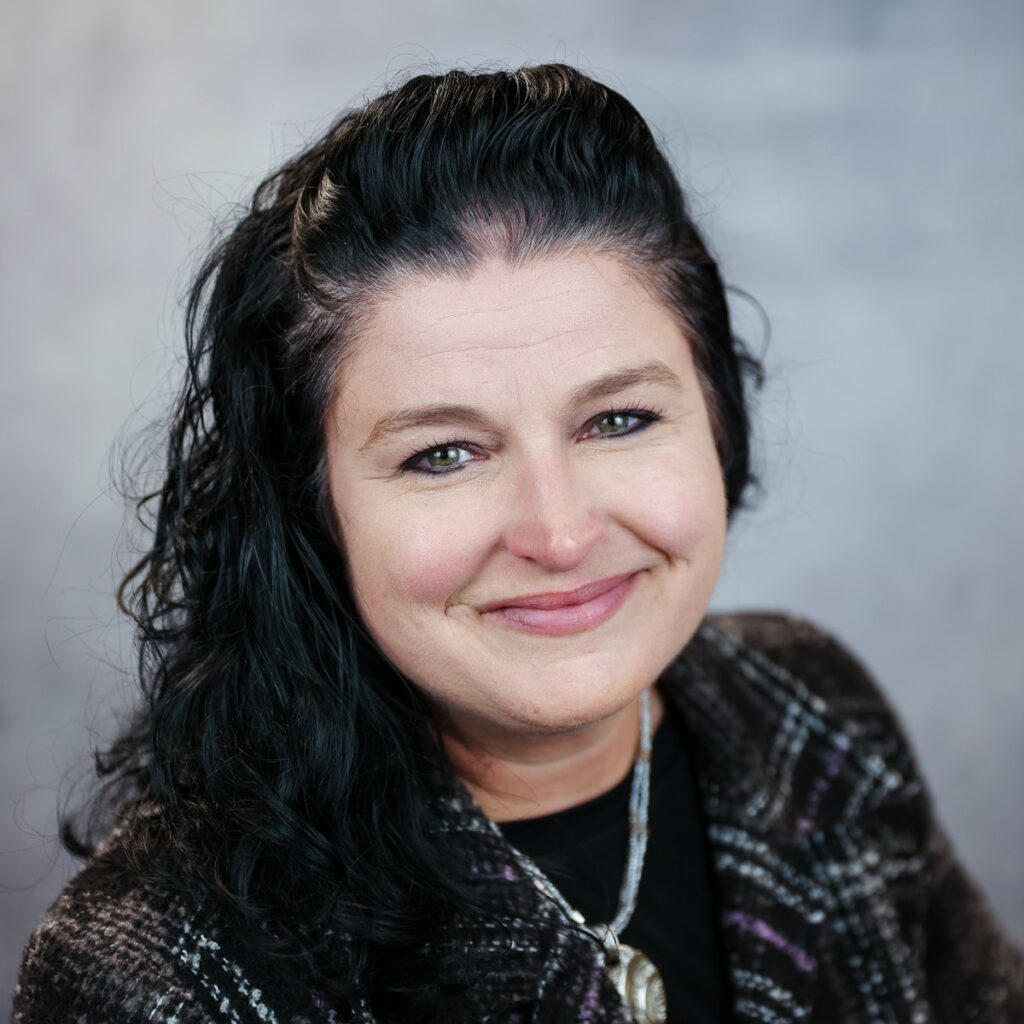 A portrait of Erin Berle, Director of employee engagement at HMC. She has dark curly hair that is half-pulled back. She wears a dark blazer and a silver necklace.