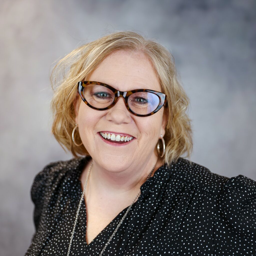 A portrait of Jen Drganc, Executive Director at HMC. She has black framed glasses and bobbed curly blond hair. She wears a black blouse. 