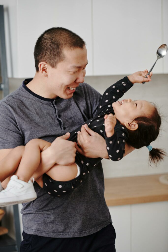 A father enrolled in IHS laughs as he holds his toddler daughter as she smiles and waves a spoon.