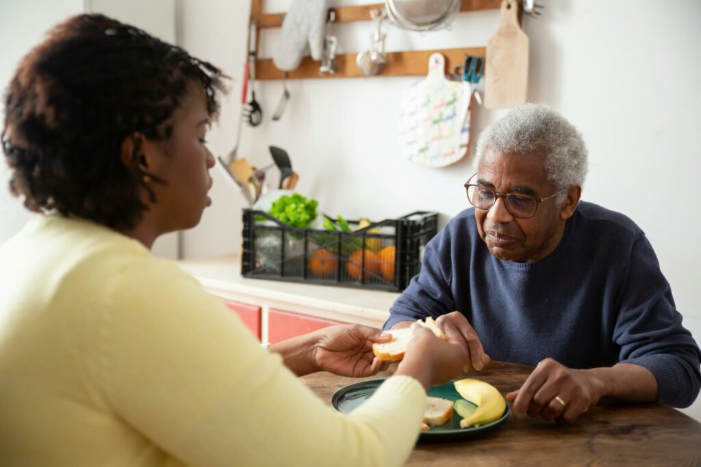 A daughter sits at the table with her elderly father, helping him with his meal. The daughter has dark skin with short curly hair. She wears a yellow sweater. The elderly man has dark skin and a dark blue sweater.